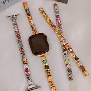 Könnte beinhalten: Drei modulare Uhrenarmbänder mit bunten Emaille- und Metallanhängern, die verschiedene Symbole, Herzen und Texte zeigen. Ein Armband ist an einem schwarzen Smartwatch-Gehäuse befestigt. Die Anhänger zeigen Blumen, ein Yin-Yang und ein Auge.