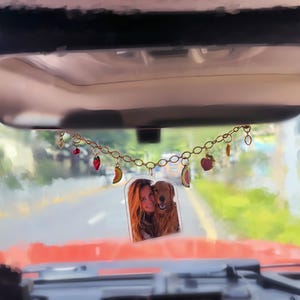 Könnte beinhalten: Ein Rückspiegel im Auto mit einer dekorativen goldfarbenen Kette, die mit Fruchtanhängern und einem transparenten Acrylfoto einer Frau und eines Golden Retrievers verziert ist. Die Anhänger zeigen Erdbeeren, Bananen und Wassermelonenstücke.