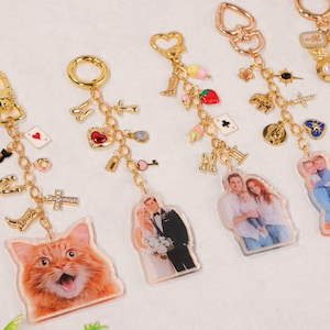 Könnte beinhalten: Eine Sammlung personalisierter Schlüsselanhänger mit goldfarbener Hardware und verschiedenen Anhängern. Einige Schlüsselanhänger zeigen Fotos von Personen, während andere Anhänger wie eine Katze, ein Herz, ein Schlüssel und Spielkarten haben. Die Anhänger sind in verschiedenen Farben.