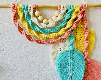 Puede incluir: Un tapiz de macramé de colores con un arcoíris de colores de hilo, incluyendo rosa, amarillo y azul. El tapiz tiene una fila de cuentas de madera y tres plumas de macramé.