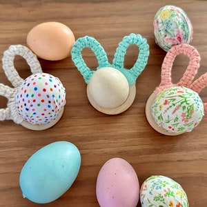 Puede incluir: Una colección de huevos de Pascua decorados con patrones coloridos y orejas de conejo de crochet. Los huevos se muestran en bases de madera.