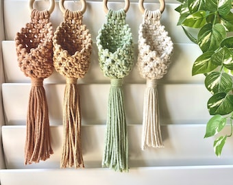 May include: Four macrame plant hangers with wooden rings and tassels. The hangers are in shades of brown, green, and white.