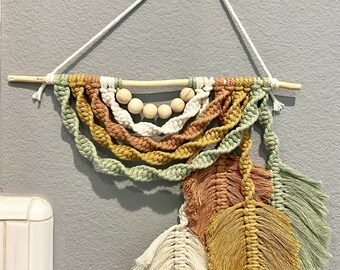 May include: A macrame wall hanging with three feathers in shades of beige, brown, and green. The hanging features a wooden dowel with a row of wooden beads.