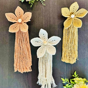 May include: Three macrame flower wall hangings with wooden beads in the center of each flower. The flowers are in shades of beige, cream, and light brown. The wall hangings are hanging on a dark brown wooden surface.