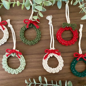 Op de afbeelding: Zes handgemaakte kerstversieringen in de vorm van kransen. De kransen zijn gemaakt van wit, groen en rood garen en zijn versierd met rode strikken.