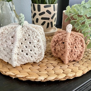 May include: Two crocheted pumpkins, one white and one light brown, sitting on a woven straw mat.