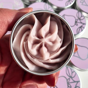 Puede incluir: Primer plano de una peque&ntilde;a lata redonda de plata con un remolino de crema rosa claro en su interior. La lata tiene un dise&ntilde;o sencillo de una rosa estilizada en la tapa.