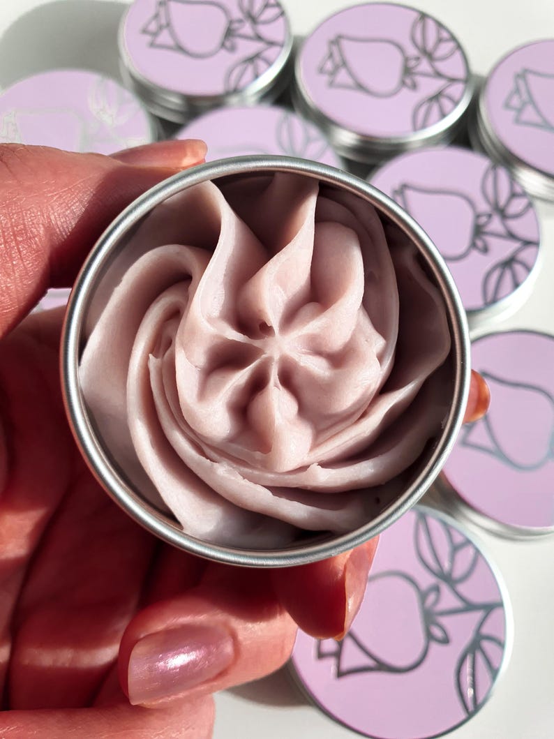 Puede incluir: Primer plano de una peque&ntilde;a lata redonda de plata con un remolino de crema rosa claro en su interior. La lata tiene un dise&ntilde;o sencillo de una rosa estilizada en la tapa.