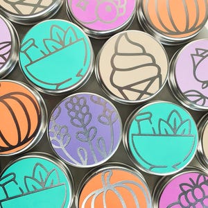 Puede incluir: Una colecci&oacute;n de 10 peque&ntilde;as latas redondas de plata con varios dise&ntilde;os coloridos. Los dise&ntilde;os incluyen una calabaza, un cono de helado, un taz&oacute;n de plantas, un racimo de bayas y flores de lavanda.