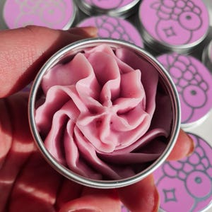 Puede incluir: Un peque&ntilde;o contenedor redondo de hojalata plateada con un remolino de crema rosa en su interior. La lata tiene una tapa plateada y est&aacute; rodeada de otras latas similares con dise&ntilde;os rosa y blanco.