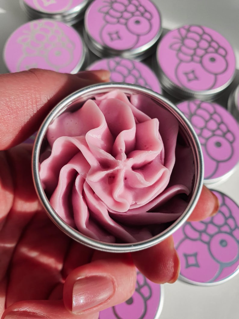 Puede incluir: Un peque&ntilde;o contenedor redondo de hojalata plateada con un remolino de crema rosa en su interior. La lata tiene una tapa plateada y est&aacute; rodeada de otras latas similares con dise&ntilde;os rosa y blanco.