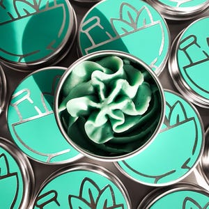 Puede incluir: Varias latas peque&ntilde;as, redondas, de plata con tapas verdes. Cada tapa tiene un dise&ntilde;o de mortero y maja con una planta que crece de &eacute;l. Una lata est&aacute; abierta, revelando un remolino de jab&oacute;n verde.