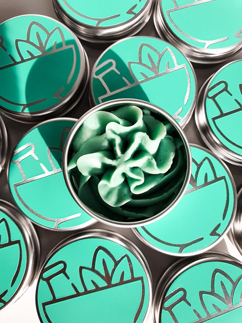 Puede incluir: Varias latas peque&ntilde;as, redondas, de plata con tapas verdes. Cada tapa tiene un dise&ntilde;o de mortero y maja con una planta que crece de &eacute;l. Una lata est&aacute; abierta, revelando un remolino de jab&oacute;n verde.