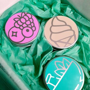 Puede incluir: Tres latas redondas de metal con tapas de colores. Las tapas presentan un cono de helado rosa con una baya en la parte superior, un cono de helado beige con un remolino de helado y un cono de helado verde azulado con una hoja verde en la parte superior.