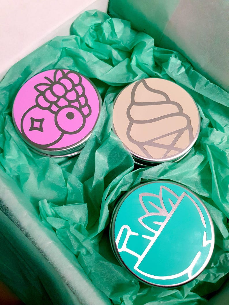 Puede incluir: Tres latas redondas de metal con tapas de colores. Las tapas presentan un cono de helado rosa con una baya en la parte superior, un cono de helado beige con un remolino de helado y un cono de helado verde azulado con una hoja verde en la parte superior.