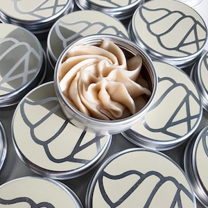 May include: Several silver tins with a swirl design on the lid. The tins are filled with a beige, whipped cream-like substance.