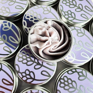 Puede incluir: Varias latas peque&ntilde;as de plata con dise&ntilde;os florales de lavanda y tapas. Una lata est&aacute; abierta y contiene un remolino de manteca corporal con aroma a lavanda p&uacute;rpura.