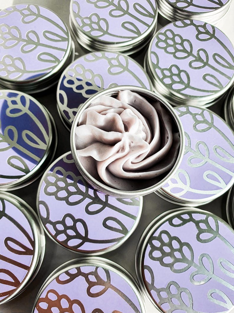 Puede incluir: Varias latas peque&ntilde;as de plata con dise&ntilde;os florales de lavanda y tapas. Una lata est&aacute; abierta y contiene un remolino de manteca corporal con aroma a lavanda p&uacute;rpura.