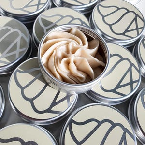 Puede incluir: Varias latas plateadas con un dise&ntilde;o de espiral en la tapa. Las latas est&aacute;n llenas de una sustancia beige que parece crema batida.