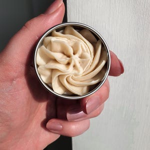 May include: A small metal tin filled with a swirl of light brown body butter.