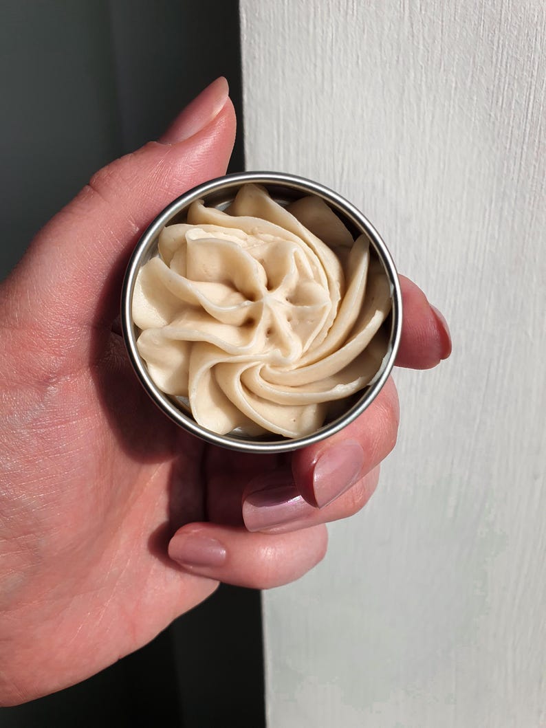 May include: A small metal tin filled with a swirl of light brown body butter.