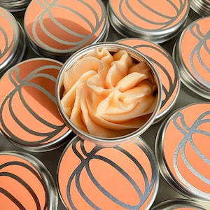 Puede incluir: Varias latas peque&ntilde;as redondas de metal con un dise&ntilde;o de calabaza en la tapa. Una lata est&aacute; abierta y contiene un remolino de mantequilla corporal de calabaza y especias de color naranja.