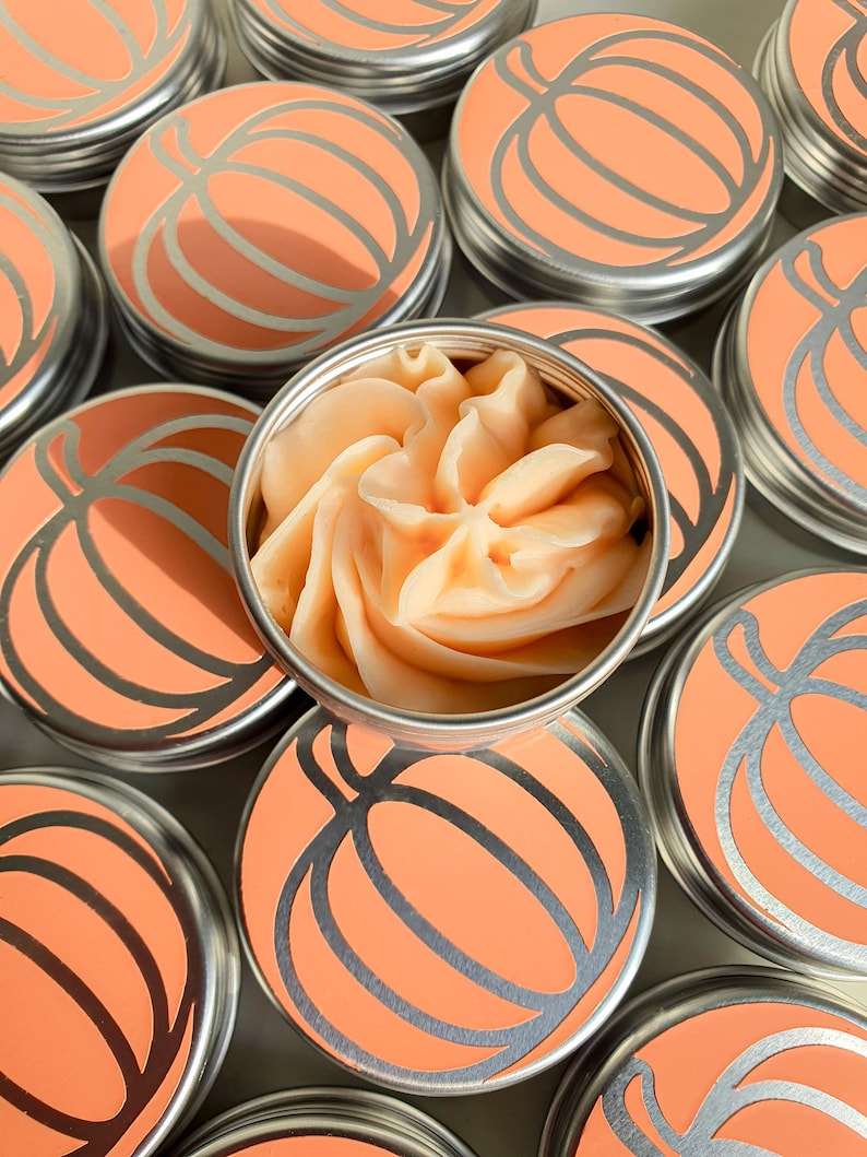 Puede incluir: Varias latas peque&ntilde;as redondas de metal con un dise&ntilde;o de calabaza en la tapa. Una lata est&aacute; abierta y contiene un remolino de mantequilla corporal de calabaza y especias de color naranja.