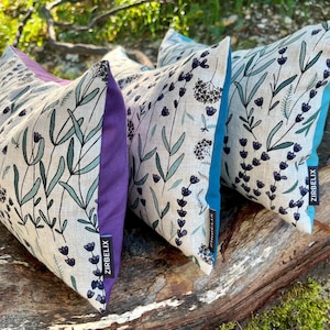 Könnte beinhalten: Drei dekorative Zierkissen mit floralen Mustern. Die Kissen haben einen hellen Hintergrund mit grünen und violetten floralen Designs. Die Seiten sind lila und türkis. Die Kissen sind mit der Marke "ZIRBELIX" versehen.