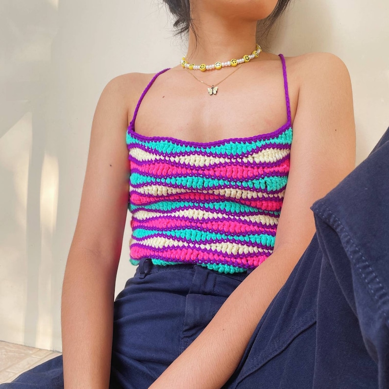 May include: A woman wearing a multi-colored striped crochet top with a purple base. The top has a thin strap design and is made with a chunky yarn. The woman is also wearing a gold butterfly necklace with a beaded chain.