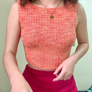 May include: A coral-coloured crochet tank top with a scoop neck. The top is made of a soft, textured yarn and has a relaxed fit.
