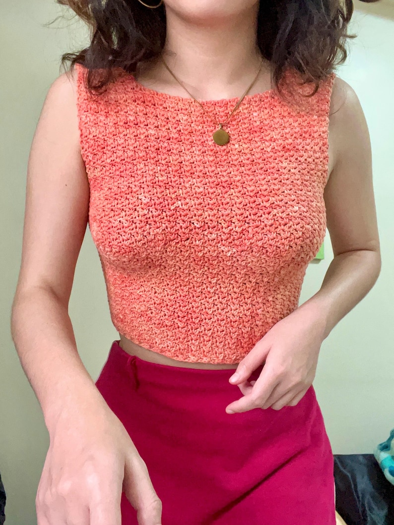 May include: A coral-coloured crochet tank top with a scoop neck. The top is made of a soft, textured yarn and has a relaxed fit.