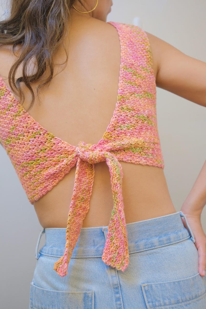 May include: A pink and green crochet top with a tie back. The top is worn over a pair of light blue jeans.