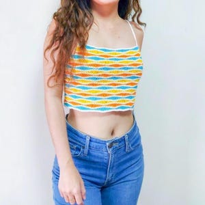 May include: A white crochet top with blue, orange, and yellow stripes. The top is sleeveless and has a cropped length. The model is wearing blue jeans.