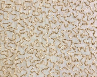 May include: A beige fabric with a gold embroidered pattern of small, interconnected loops. The fabric has a subtle texture and a slightly faded appearance.