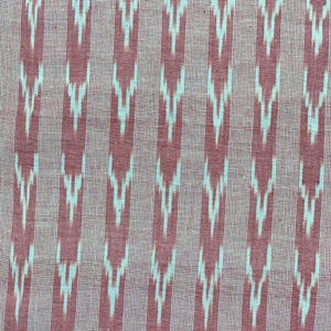May include: Pink and white fabric with a repeating ikat pattern. The fabric has a subtle texture and a slightly faded appearance.