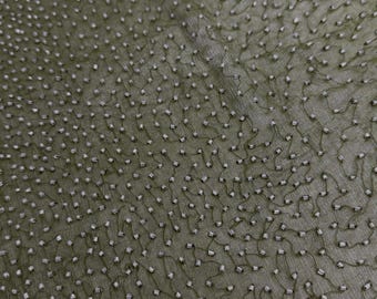 May include: Close-up of olive green fabric with a delicate pattern of small, square, silver-colored beads. The beads are connected by thin, dark green threads, creating a textured surface. The fabric has a subtle sheen.