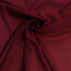 May include: A deep burgundy fabric with a smooth, matte texture, arranged in a central swirl pattern that creates soft, flowing folds and shadows across the surface, highlighting the consistent dark red color and the drape of the material.