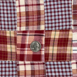 May include: A patchwork fabric with a red, white, and blue plaid pattern. The fabric is made up of different squares of fabric sewn together. A silver quarter is placed on the fabric for scale.