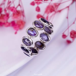 May include: A silver ring with a band of oval-shaped purple gemstones. The ring is set on a white surface.