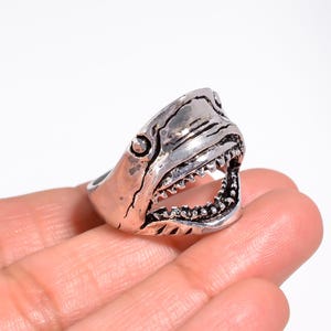 May include: A silver-toned metal ring shaped like a shark head with an open mouth, showing detailed teeth and a textured, weathered finish. The design features a prominent eye and gill slits, creating a realistic representation of a shark.