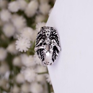 May include: A silver-toned metal ring featuring a detailed, sculpted figure with flowing hair and arms crossed over the chest. The design has an oxidized finish that highlights the intricate textures and facial features of the central character.