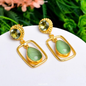 Könnte beinhalten: Goldene Ohrringe mit grünen Edelstein-Tropfen. Die Ohrringe haben einen quadratischen Rahmen mit einem tropfenförmigen Stein im Inneren. Der obere Teil der Ohrringe hat einen kleineren runden grünen Stein.