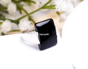 Puede incluir: Un anillo de plata con una piedra preciosa negra de forma cuadrada. El anillo está colocado sobre una superficie blanca.