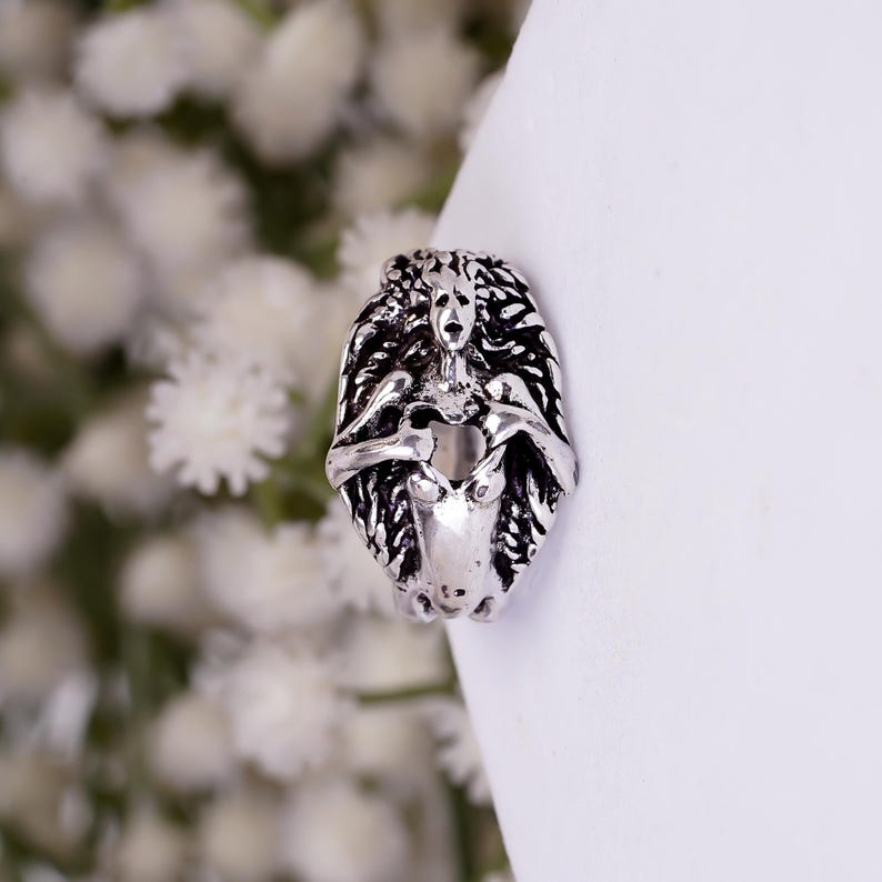 May include: A silver-toned metal ring featuring a detailed, sculpted figure with flowing hair and arms crossed over the chest. The design has an oxidized finish that highlights the intricate textures and facial features of the central character.