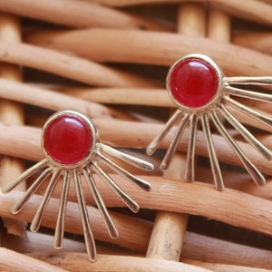 Può includere: Un paio di orecchini d'argento con accenti di pietre preziose rosse. Ogni orecchino presenta una pietra rossa rotonda circondata da raggi argentati.