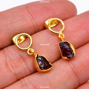 Może przedstawiać: Para złotych kolczyków z wiszącym, ciemnoczerwonym kamieniem szlachetnym. Kolczyki mają okrągły obręcz na górze i mały, wiszący kamień szlachetny.