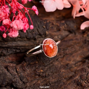 May include: A silver ring with a red and orange sunstone gemstone. The ring is on a wooden surface.