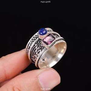 Può includere: Un anello d'argento con una montatura di pietre preziose blu e viola. L'anello presenta un design intricato con una fascia testurizzata.