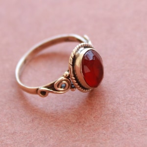 Puede incluir: Un anillo de plata con una piedra roja ovalada engastada en un engaste de plata decorativo. El anillo tiene un diseño delicado y ornamentado con un patrón de espiral en un lado.
