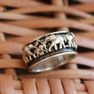 May include: A silver ring with a band of elephants walking around the ring. The elephants are in a line and are facing the same direction. The ring is on a woven brown background.